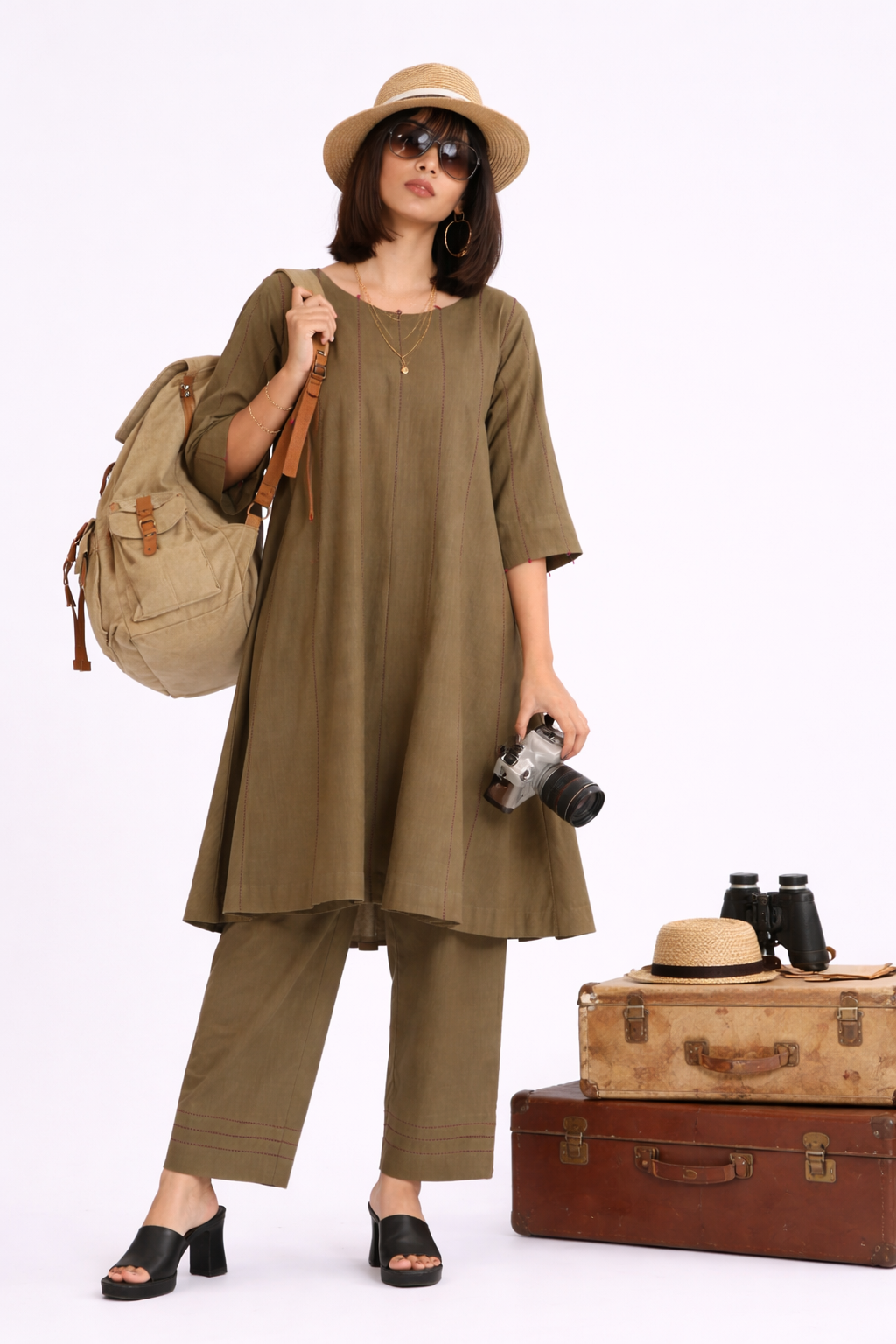 Woman in olive green outfit with hat, backpack, and camera, standing next to vintage suitcase and binoculars on a white background.