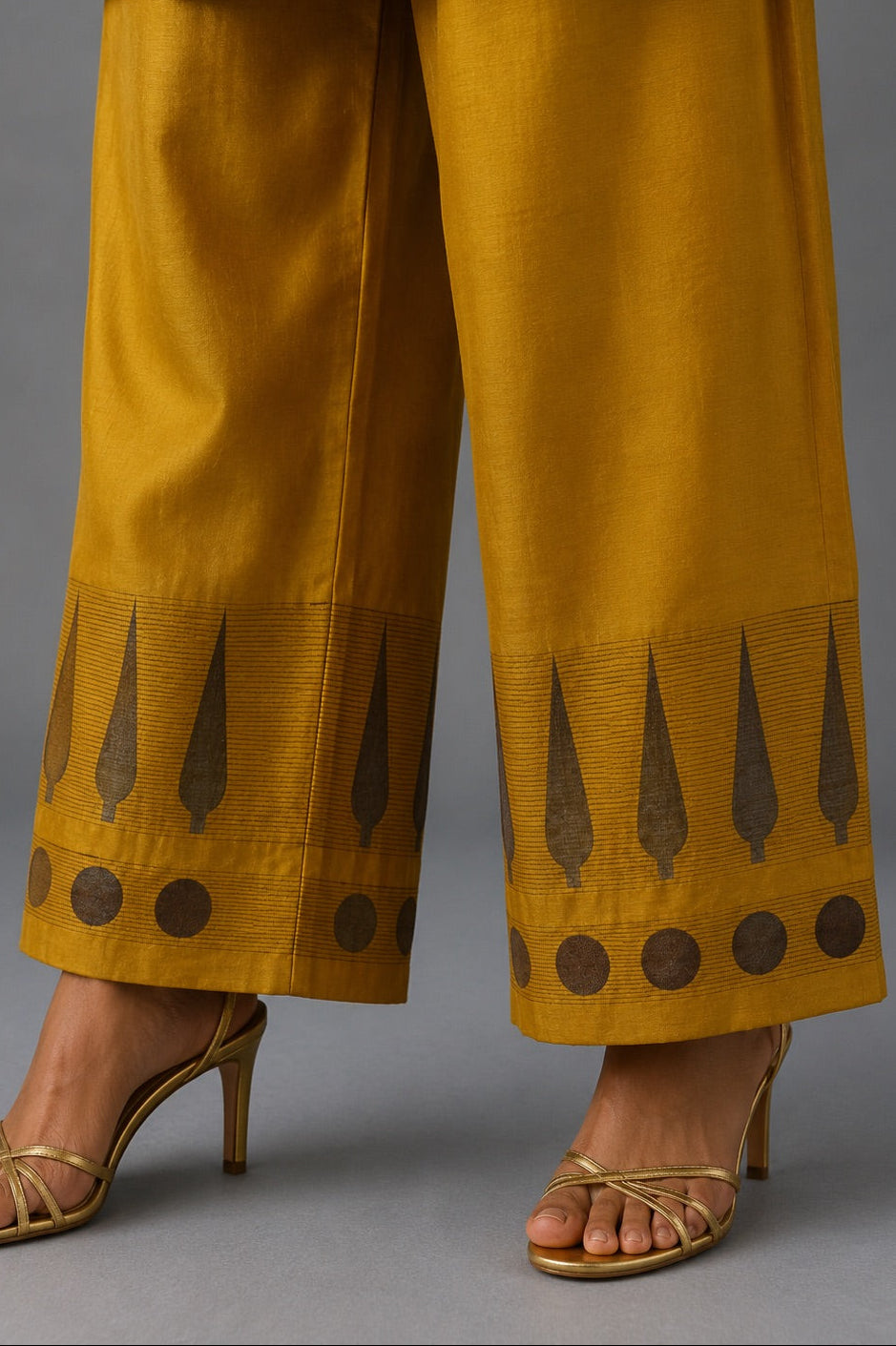 Close-up of a person wearing mustard yellow wide-leg pants with dark brown geometric border patterns near the hems, paired with gold heeled sandals against a gray studio background.