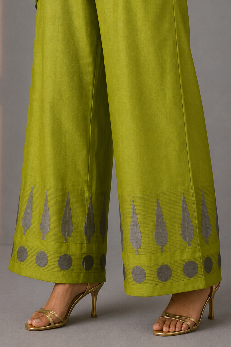 A person wearing bright olive green wide-leg pants with gray geometric patterns near the hems, paired with metallic gold heeled sandals against a gray studio background.